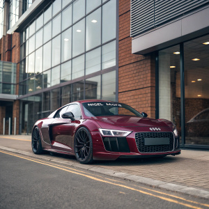 Digital art of sleek, maroon Audi R8 parked outside modern office building. High-performance sports car with glossy paint and bold grille, set against backdrop of glass and brick facade. Nigel Mora logo on car