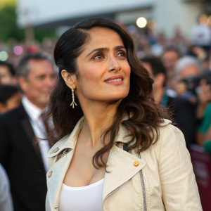 Salma Hayek in a casual outfit, attending a charity gala. The event is outdoors with a bustling crowd and vibrant atmosphere. Close-up portrait style.