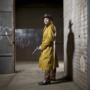 Karl Heisenberg, in trench coat, boots, and fedora, holding a vintage pistol, standing in a dimly lit alleyway with graffiti-covered walls.