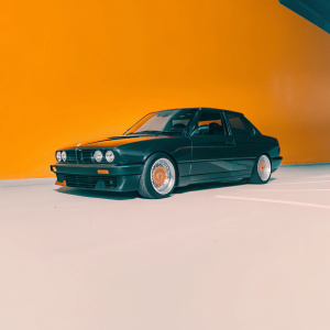 A sleek black BMW M3 E30 parked on a bustling street at night. The car features a custom front bumper, sharp headlights with distinctive markings, and large tires with red accents. Dark background highlights the vehicle's low stance and aerodynamic design.