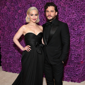 Emilia Clarke and Kit Harington, in elegant attire, at the Met Gala. Emilia in a floor-length black gown with a bow detail. Kit in a tuxedo. They pose formally against a purple floral backdrop.
