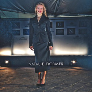 Natalie Dormer, smiling confidently in a black skirt suit, at a corporate event. The venue features a sleek white backdrop adorned with company logos and text.
