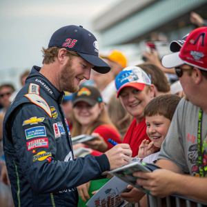 Dale Earnhardt Jr. signing autographs at NASCAR event. Fans in team jerseys and hats surround him.