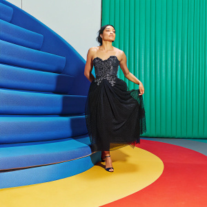 Michelle Rodriguez, wearing a stunning black dress with intricate details, at a Hollywood gala. The dress features a fitted bodice with embellishments and a flowing skirt. She stands on a blue-patterned stage. High-resolution photo with blue and gray textured backdrop.