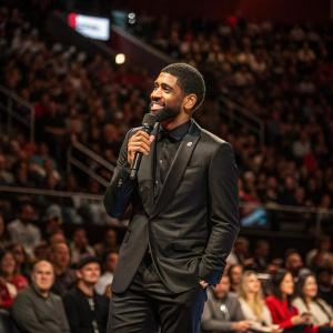 Kyrie Irving, wearing a sleek black suit, stands in front of a crowded auditorium at a sports conference. He's holding a microphone, addressing the audience.