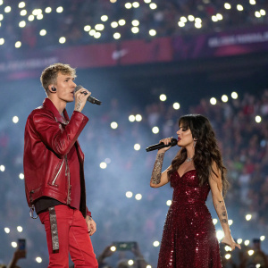 Machine Gun Kelly and Camila Cabello mid-performance at a packed stadium. MGK in red leather jacket, Camila in maroon sequined dress. Flashing camera lights, fans holding up phones