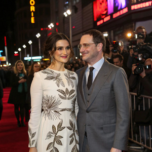 Rachel Weisz and Darren Aronofsky at a bustling gala event. Rachel is in a stunning white dress with intricate brown patterns. Darren is in a tailored gray suit with a dark tie. They are surrounded by flashing cameras and crowd in a cityscape backdrop with neon lights.