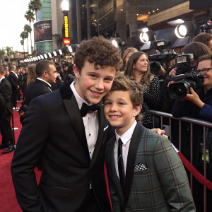 Nolan Gould and an unknown young man posing together at a vibrant Hollywood event. They are surrounded by flashing cameras and paparazzi.