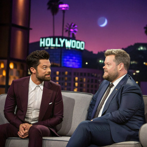 Dominic Cooper and James Corden on a gray couch during a live talk show. Purplish studio lights, Hollywood sign visible in the background.