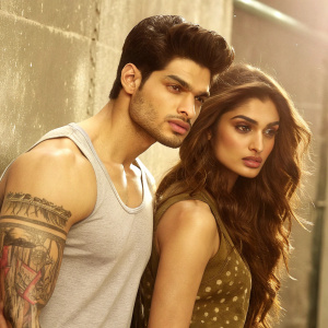 Sooraj Pancholi and Athiya Shetty, stars of Hero 2015, pose for promotional shoot. Sooraj with muscular build, tattooed arm, Athiya with serious expression. Grungy textured background, warm golden tone.