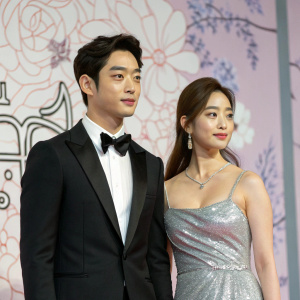 Seo Kang-Joon and Kim Q-Ri, starring in 'Sly and Single Again,' appear at a glamorous awards show. Seo Kang-Joon in a sleek black tuxedo, Kim Q-Ri in a shimmering silver gown. The backdrop features intricate floral patterns in pastel shades.