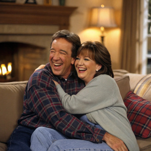 Tim Allen and Patricia Richardson, former 'Home Improvement' stars, hugging and laughing. They are sitting on a plush couch in a cozy living room setting.