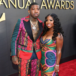 YFN Lucci and Reginae Carter at a gala event. YFN Lucci in a bold, multi-colored suit, with detailed tattoos on his chest. Reginae Carter wearing a colorful dress. Red carpet backdrop with gold letters reading 'Annual Awards'