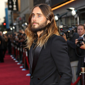 Jared Leto, with a beard and long wavy hair, wearing a black suit. Attending the premiere of his new film, on the bustling Hollywood red carpet.