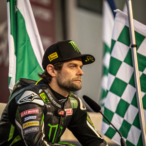 Cal Crutchlow, serious expression, in racing gear. Sitting in front of green and white checkered flags. Surrounded by microphones.