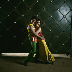 Shah Rukh Khan and Kajol, both with dark skin, at a fun photo shoot. Wearing vibrant outfits, they share a playful hug. Silver and black backdrop.