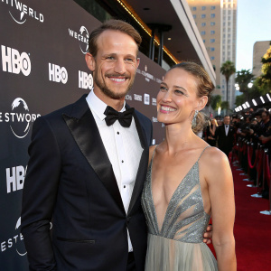 Gustaf Skarsgård and Caroline Sjöstrand attending a Westworld premiere. He's in a tuxedo, she's in a stunning gown. Both have warm smiles. Red carpet, flashing cameras, and HBO branding in the background.