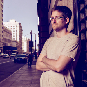 Edward Snowden, a white male with a stern look, short brown hair, and glasses, standing in front of a busy city street. He is wearing a casual t-shirt.