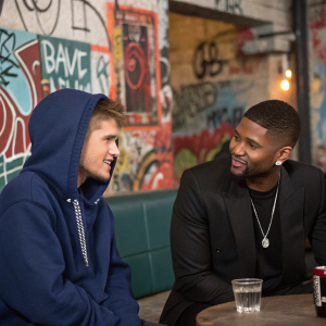 Justin Bieber and Usher at a casual hangout. Justin wears a navy blue hoodie, while Usher is in a black blazer. They're surrounded by graffiti-covered walls and urban street art.