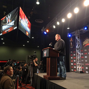 Brock Lesnar at a WWE press conference, dressed in casual attire. Standing behind a podium under bright lights. A large screen displays WWE graphics and advertisements. The room is filled with reporters and photographers.