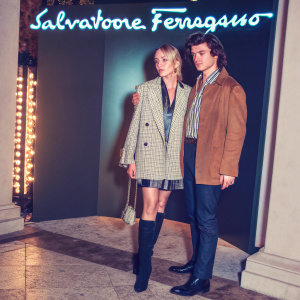 Maika Monroe and Joe Keery at the Salvatore Ferragamo fashion show in Milan. Monroe wears a chic plaid jacket with black boots. Keery is in a brown jacket and striped shirt. The backdrop features the Salvatore Ferragamo logo.