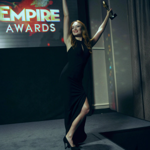 Sophie Turner posing confidently at the Empire Awards press room, adorned in a sleek black dress. Vibrant backdrop with Empire Awards logos.