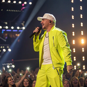 Pitbull at the 2023 Billboard Music Awards, wearing a neon yellow jacket and a white cap. Poses confidently on stage amidst flashing lights and a crowd of fans.
