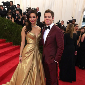 Chloe Bridges and Adam DeVine at the Met Gala. Chloe in a gold floor-length gown, Adam in a maroon tuxedo. Crowded red carpet scene.