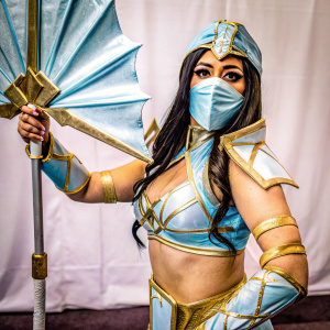 Kitana, a fictional female ninja character from Mortal Kombat, dressed in a blue and gold armored outfit. Long dark hair flowing behind her. She holds a weapon with confidence against a plain white backdrop.