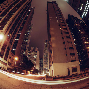 A panoramic nighttime view of Hong Kong's skyline. Towering skyscrapers of various heights, adorned with glowing windows and neon lights, stretching upwards against the dark sky. Focused low-angle shot emphasizing architectural details and moving light trails. Urban environment bustling with life.