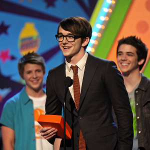 Drake Bell, actor, on stage at Nickelodeon's Kids' Choice Awards 2008, receiving an award. He wears a dark suit, brown tie, glasses. Two other individuals, smiling, stand nearby. The colorful, dynamic stage behind him provides a vivid backdrop.