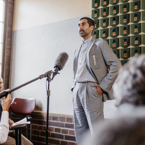 Pep Guardiola, in a casual suit, addressing the media during a press conference. The backdrop is a green wall with team badges.