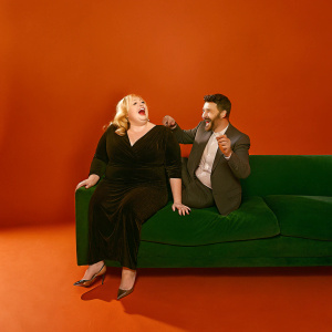 Rebel Wilson and Jimmy Kimmel laughing at a late-night talk show set. Wilson in a black dress, Kimmel in a suit.