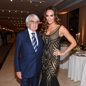 Bernie Ecclestone in a navy blue suit and striped tie. Tamara Ecclestone in a black gown with intricate detailing. They are at a formal event with elegant indoor lighting and tables set up.