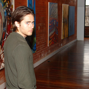 Jared Leto, intense gaze, in a modern art gallery. Wearing a casual, dark green sweater. Brightly lit, abstract paintings in the background.