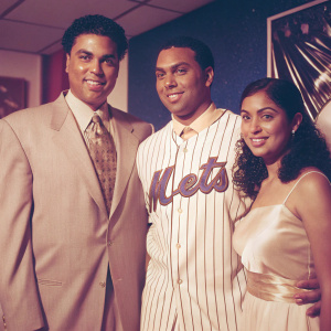Carlos Beltran in a Mets jersey stands alongside Omar Minaya, formally dressed, and his wife Jessica. The backdrop features blue and vibrant Mets logos. All smiling for the camera at an indoor event.