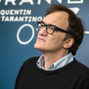 Quentin Tarantino, the renowned film director, in a casual outfit. Black sweater, striped collar, black-rimmed glasses. His expression is somber. Behind him, a vast expanse of deep blue with white text