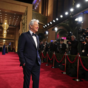 Richard Gere, in a formal navy suit, at a gala event. Lavish red carpet with flashing cameras.