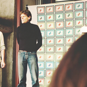 Close-up of Willem Dafoe at the Tech Innovators Conference. He sports a casual black turtleneck and jeans, standing in front of a vibrant, colorful backdrop with startup logos.
