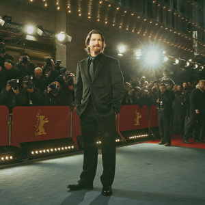 Christian Bale, Caucasian male, wearing a black suit with a beard, standing on a red carpet at Berlinale International Film Festival. Surrounded by photographers and excited fans.