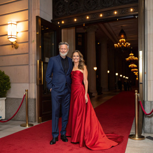 Bill Pullman and Tamara Hurwitz attending a charity gala. Bill in a navy suit, Tamara in a flowing red gown. Luxurious red carpet entrance.