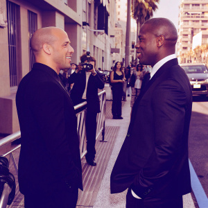 Vin Diesel and Tyrese Gibson, dressed in black suits, stand on a busy street during the Fast & Furious 6 promotional tour.