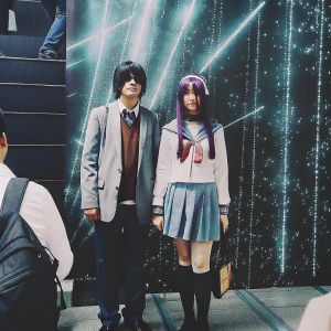 Haruhi Suzumiya, long purple hair, determined look, standing in front of a bright, animated backdrop filled with sparkles and neon colors. Her companion, Koizumi Itsuki, stands beside her with a calm demeanor. They are at a vibrant anime convention.