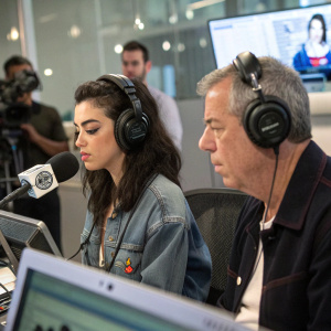 Charli XCX and Elvis Duran in the studio. Both wearing headphones. Microphones and camera visible. Modern recording setup.