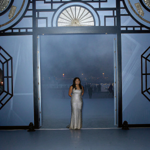 Jenna Ushkowitz, an Asian actress, in a shimmering gown. Standing in front of a dimly lit, artistic backdrop of black and gold. The setting is an indoor event with a bustling crowd in the background.