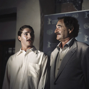 Shia LaBeouf, surprised expression, stands beside Oliver Stone. Stone has a mustache and looks kind. Event backdrop with logos.