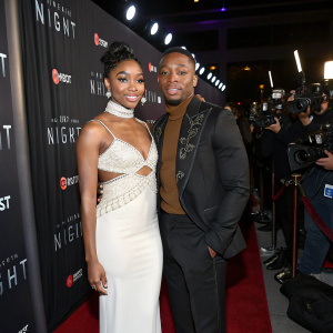 LaToya Tonodeo and Arlen Escarpeta attend the Hollywood premiere of 'I Am The Night'. LaToya in a form-fitting white gown with intricate beading. Arlen in a sophisticated black and brown ensemble. The backdrop is a sleek black curtain with glowing sponsor logos. A sea of flashing cameras and intrigued onlookers surround them.