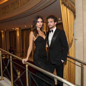 Emily Ratajkowski and Alex Sapir at a stylish gala. They stand by a sleek railing, against a backdrop of luxurious drapes.