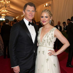 Chef Bobby Flay and actress Helene Yorke at a glamorous charity gala. Both are dressed in formal attire: Bobby in a sleek black tuxedo, Helene in a flowing white gown with delicate embroidery.