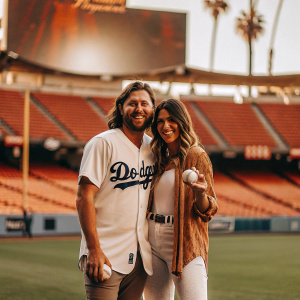 Caucasian duo Misty May-Treanor and Matt Treanor posing on Dodger Stadium's lush green field. Matt in Dodgers jersey, Misty holding baseball. Stadium's towering stands and massive screen backdrop. Palm trees in the distance, capturing a candid moment.